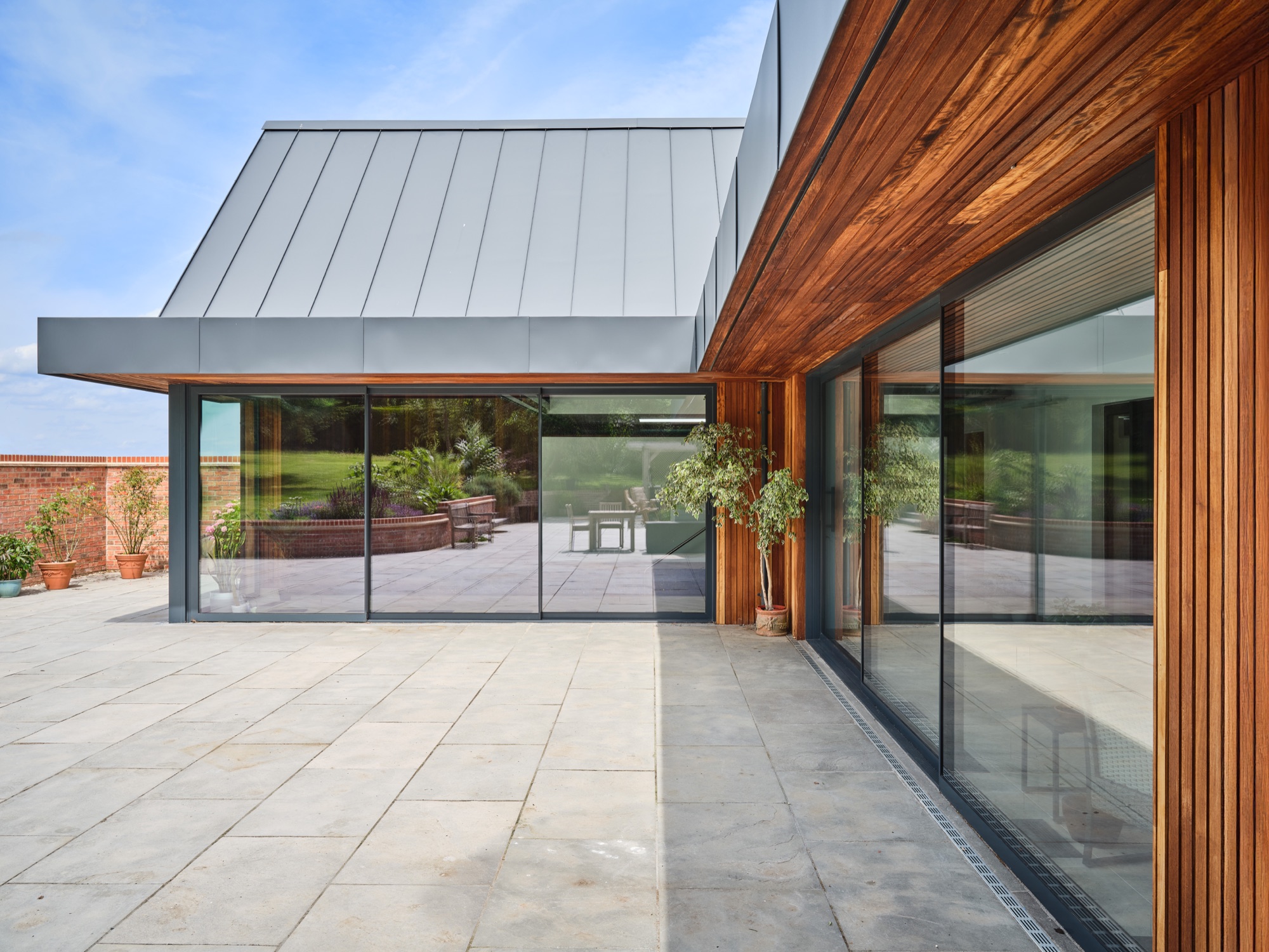The terrace running along the full glazed front of the pool hall &mdash; standing-seam zinc roof, full-height sliding glazing, open countryside beyond