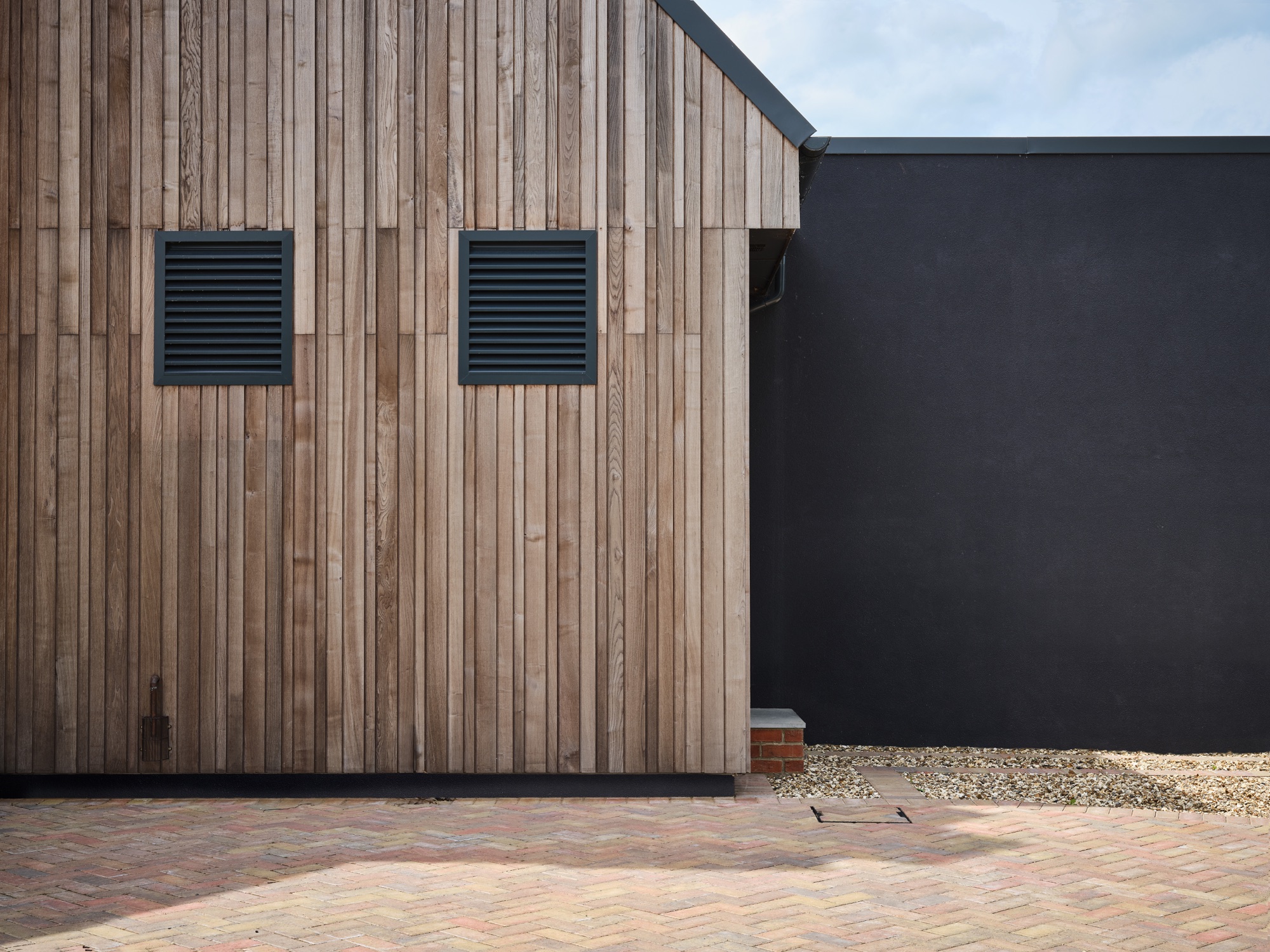 Graphic view of the timber cladding against the dark render wall &mdash; vertical boards, louvred ventilation panels, herringbone brick foreground