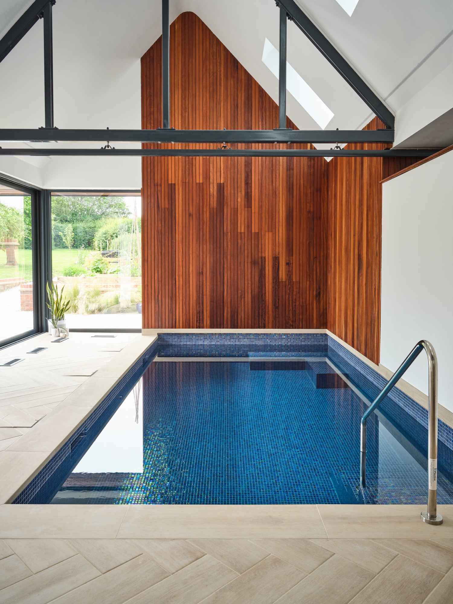 The pool from the far end &mdash; blue mosaic tile, full-height Iroko feature wall, expressed steel trusses and roof skylights
