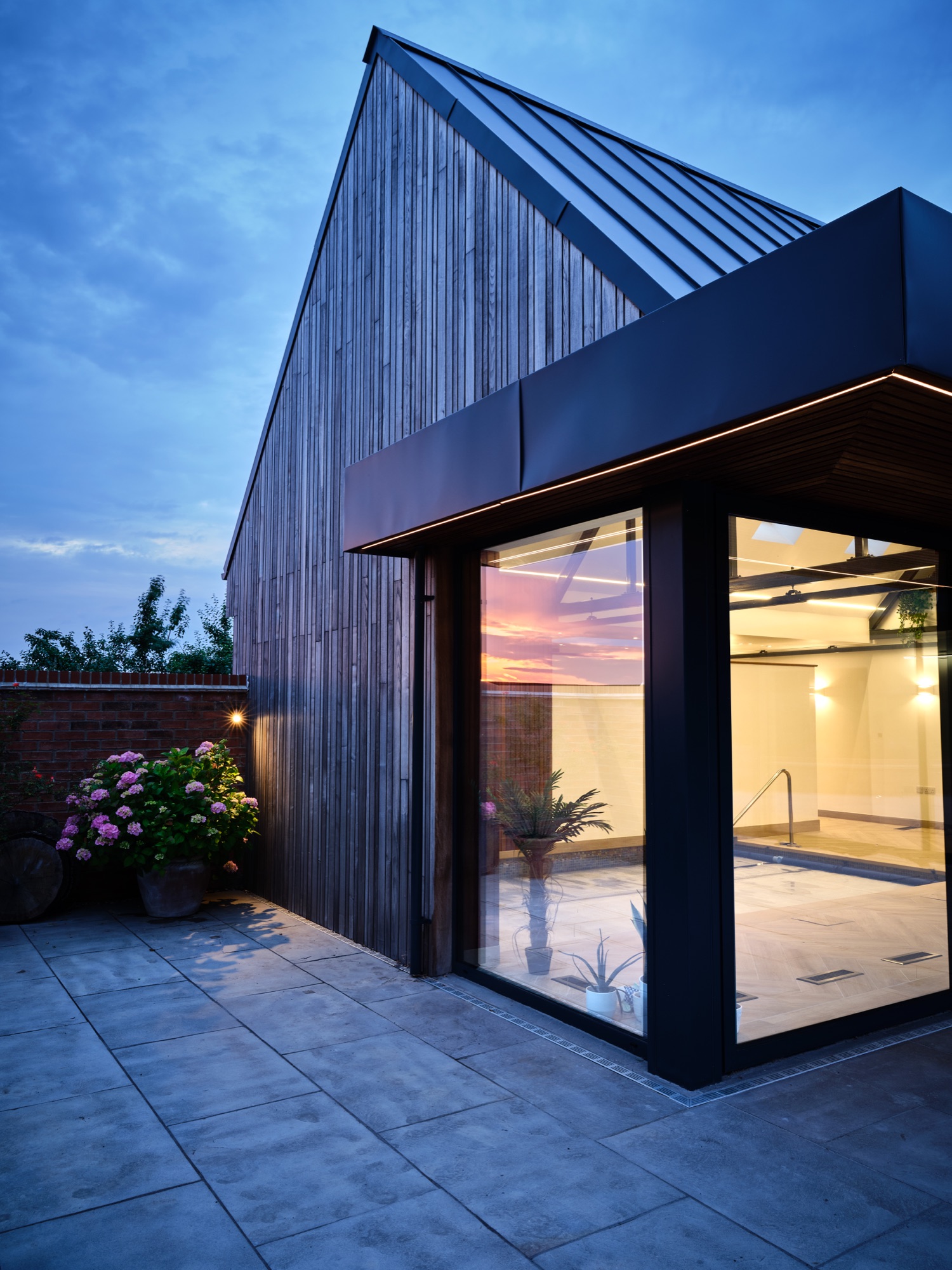 The gable end at dusk &mdash; timber cladding silhouetted, sunset colours reflected in the full-height glazing