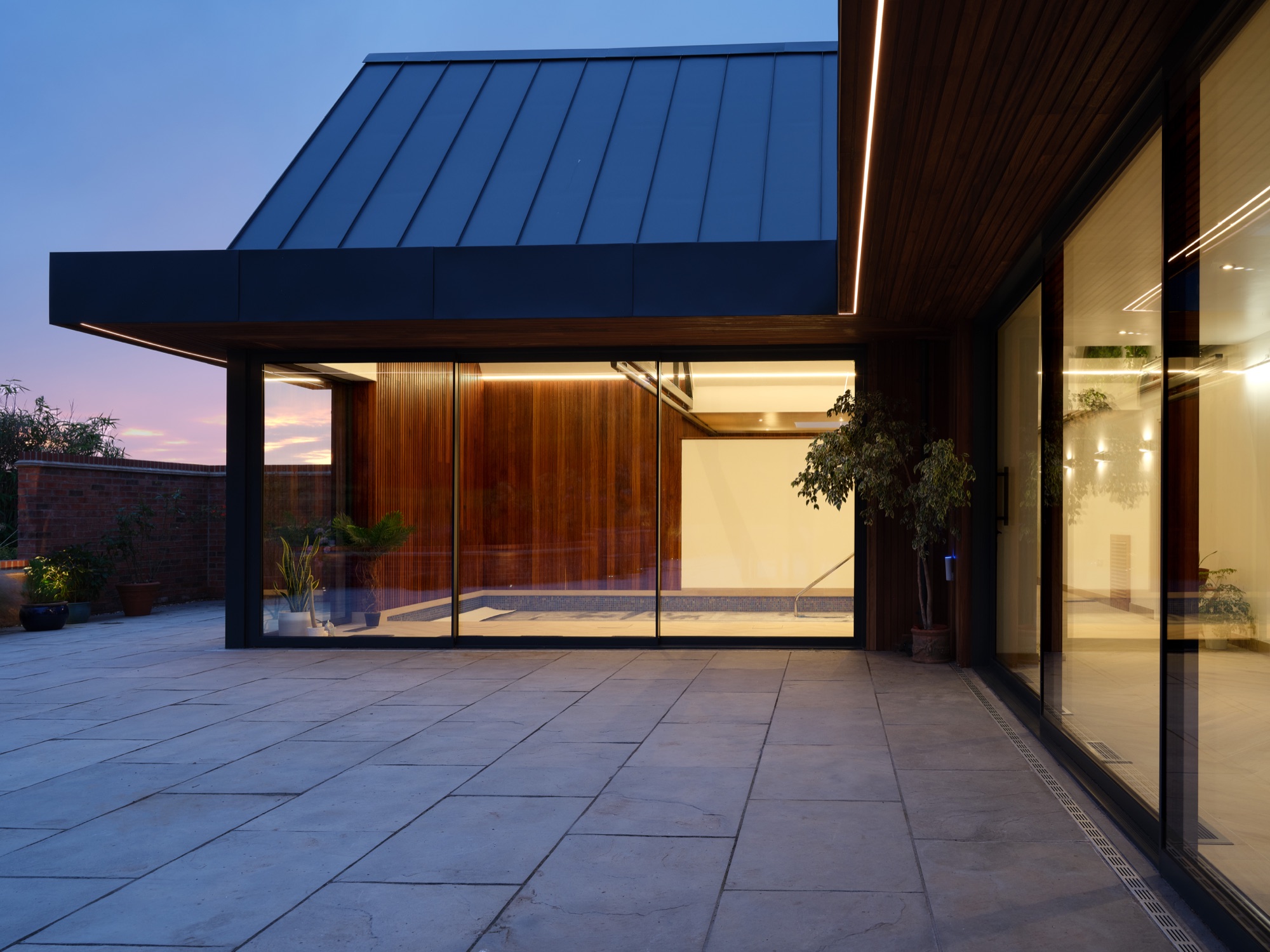 Dusk from the courtyard &mdash; the pool hall glowing from within, warm light through the full-height glazing, LED strip in the Iroko soffit