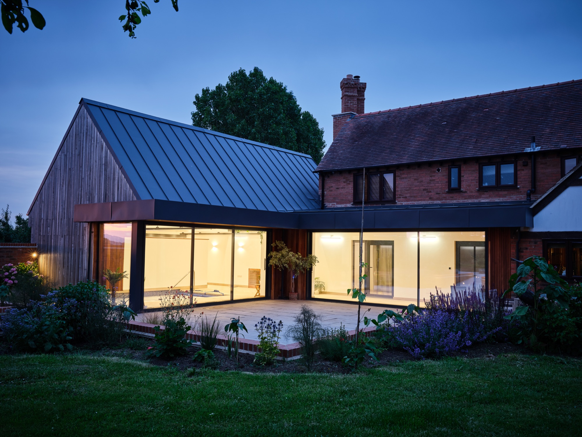 The full composition at dusk from the garden &mdash; the new pool hall and the original house together, warmly lit against the evening sky